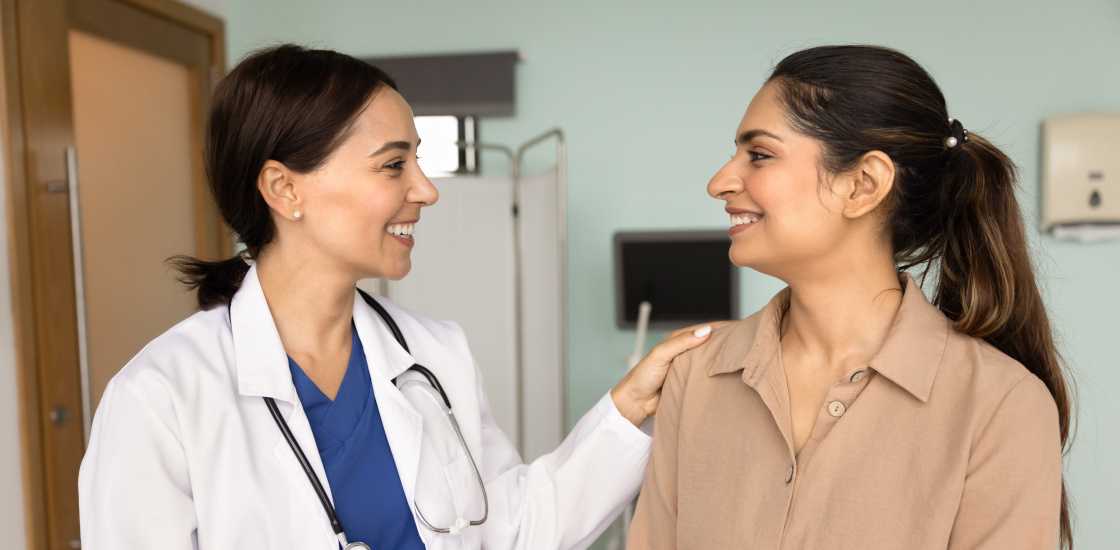 A female patient sitting beside a female physician, both smiling, as physician gives patient calming reassurance.