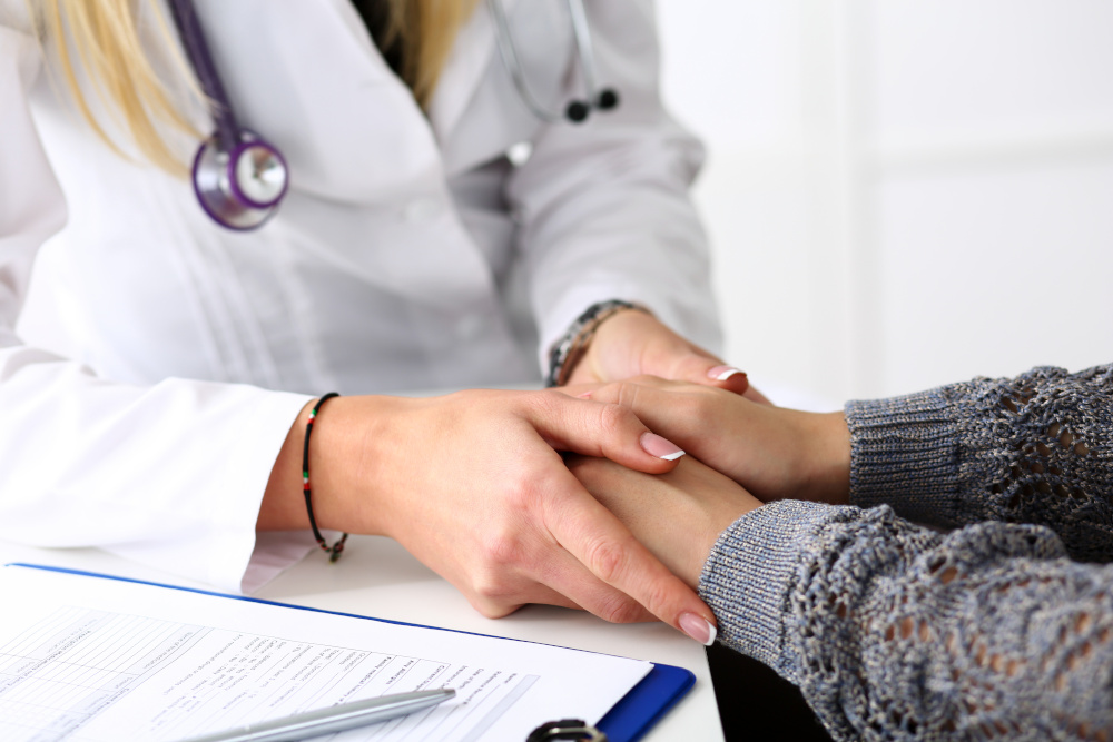 A closeup of a female doctor calmy holding the hands of a female patient in a comforting manner, a clipboard of patient intake questions on the table beside them.