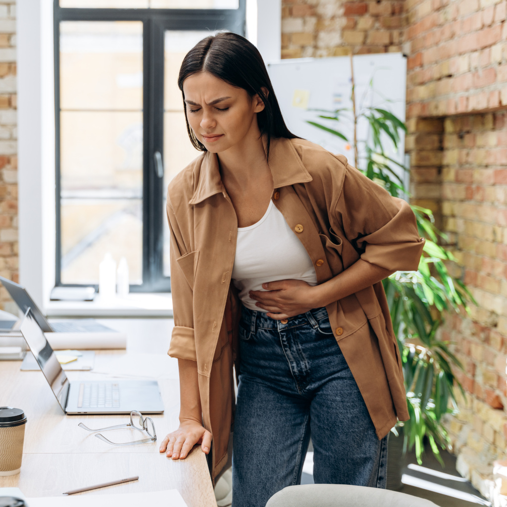 A woman stands in a sunlit office, hand on her abdomen clearly in pain.