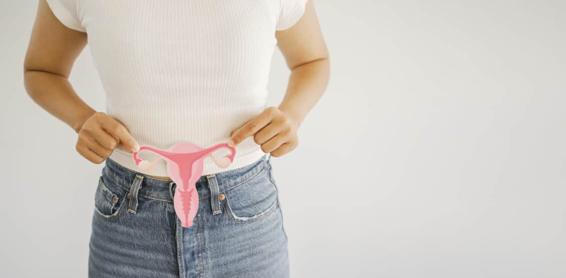 A woman standing in front of a white backdrop holding a cutout of a uterus in front of her pelvis.