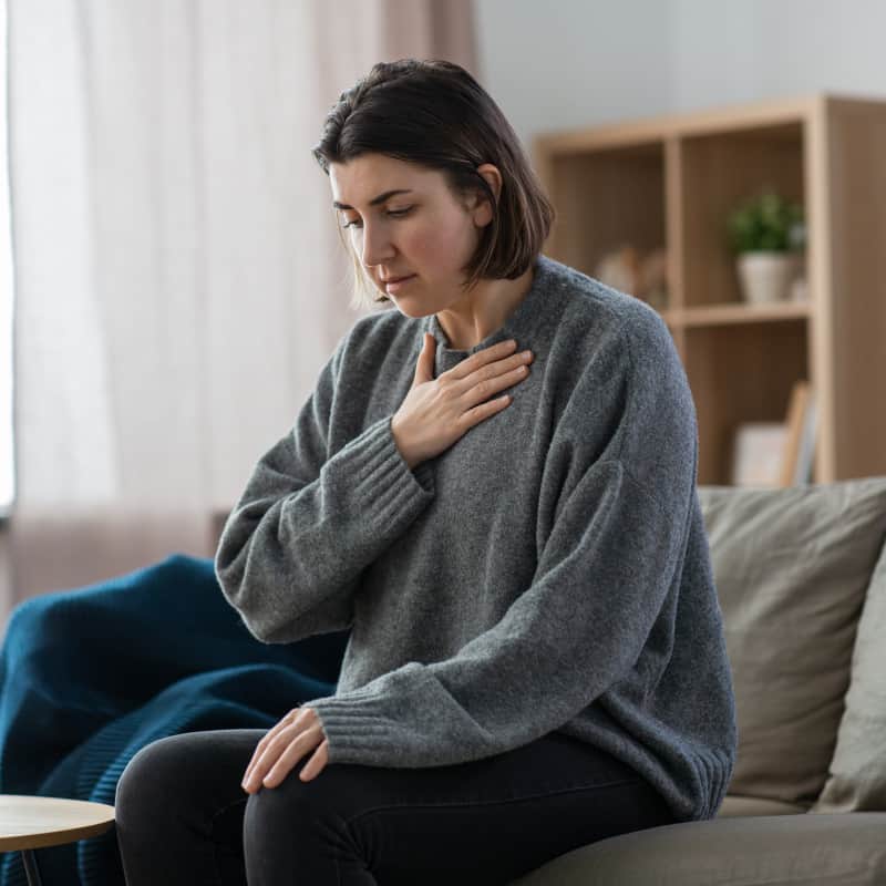 A woman sitting on a couch with her hand to her upper chest with a look of discomfort