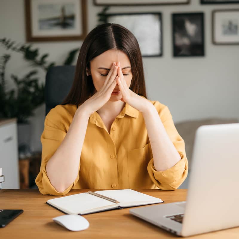 A woman with her hands over her face sitting at her desk looking exhausted