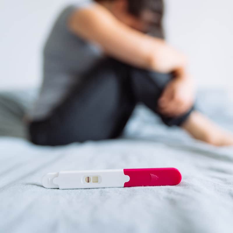 A woman in the background sitting on her bed looking somber, with a negative pregnancy test in the foreground