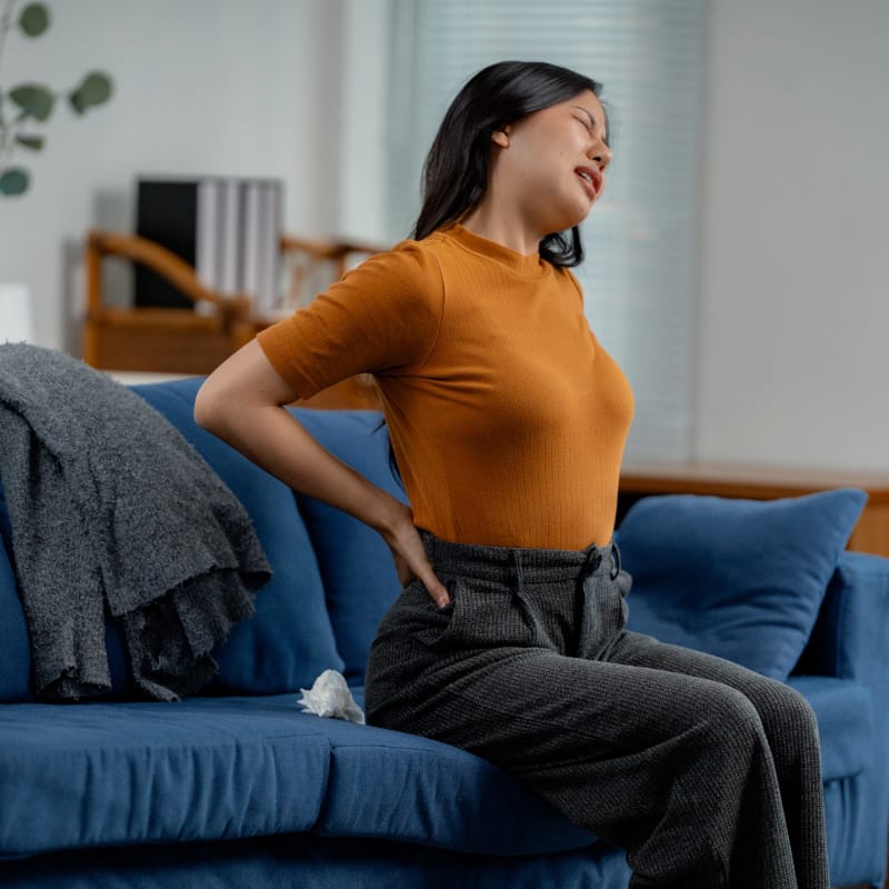 A woman sitting down arching slight backwards with her hands on her back with a look of discomfort on her face