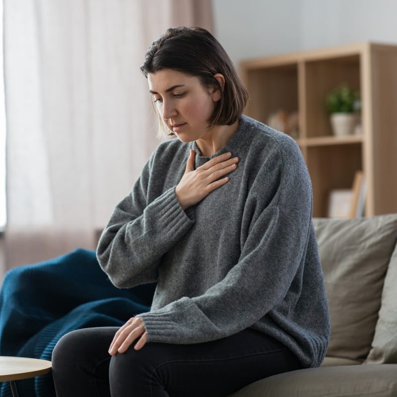 A woman sitting on a couch with her hand to her upper chest with a look of discomfort