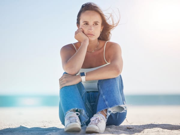A woman sitting on the beach, ocean in the background, looking towards camera with somber expression
