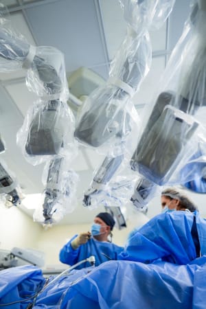 A male and female physician in scrubs performing surgery in the OR with 3 robotic arms from the da Vinci robotic surgery machine in the foreground