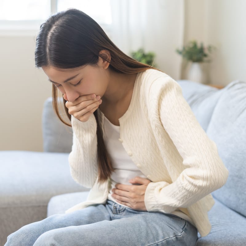 A young Asian female sitting on a couch with her hand over her mouth and other hand on her stomach looking sick