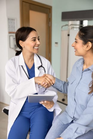 A young female doctor shakes the hand of a young female patient, both smiling.