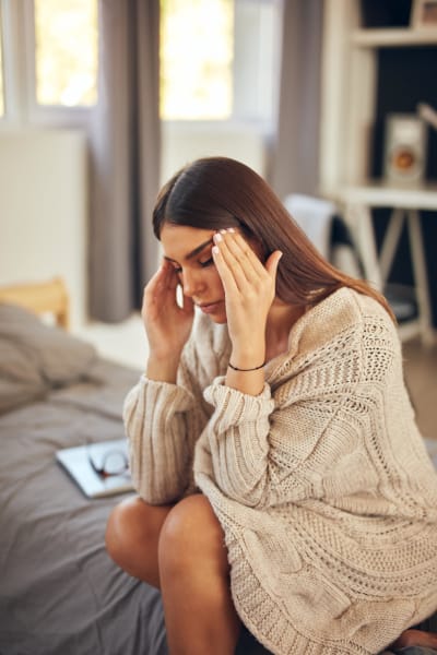 A female sitting on her bed rubbing her temples, eyes closed, clearly uncomfortable