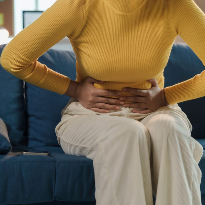 A woman sitting on a couch facing toward the camera with her hands over her lower abdomen hunched over in pain