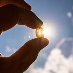 A hand holding up a vitamin D supplement pill obscuring the sun.