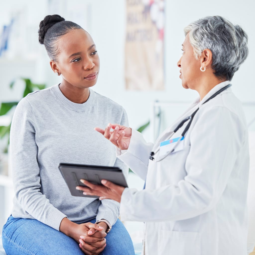A black woman sitting on an exam table speaking with her female physician about her symptoms.