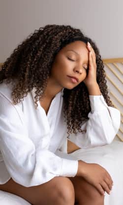 A closeup of a distressed woman sitting on the edge of her bed, hand over part of her face with eyes closed