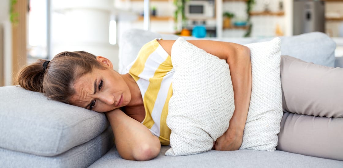 A young female laying down on a couch, in discomfort, holding a pillow tightly against her abdomen.